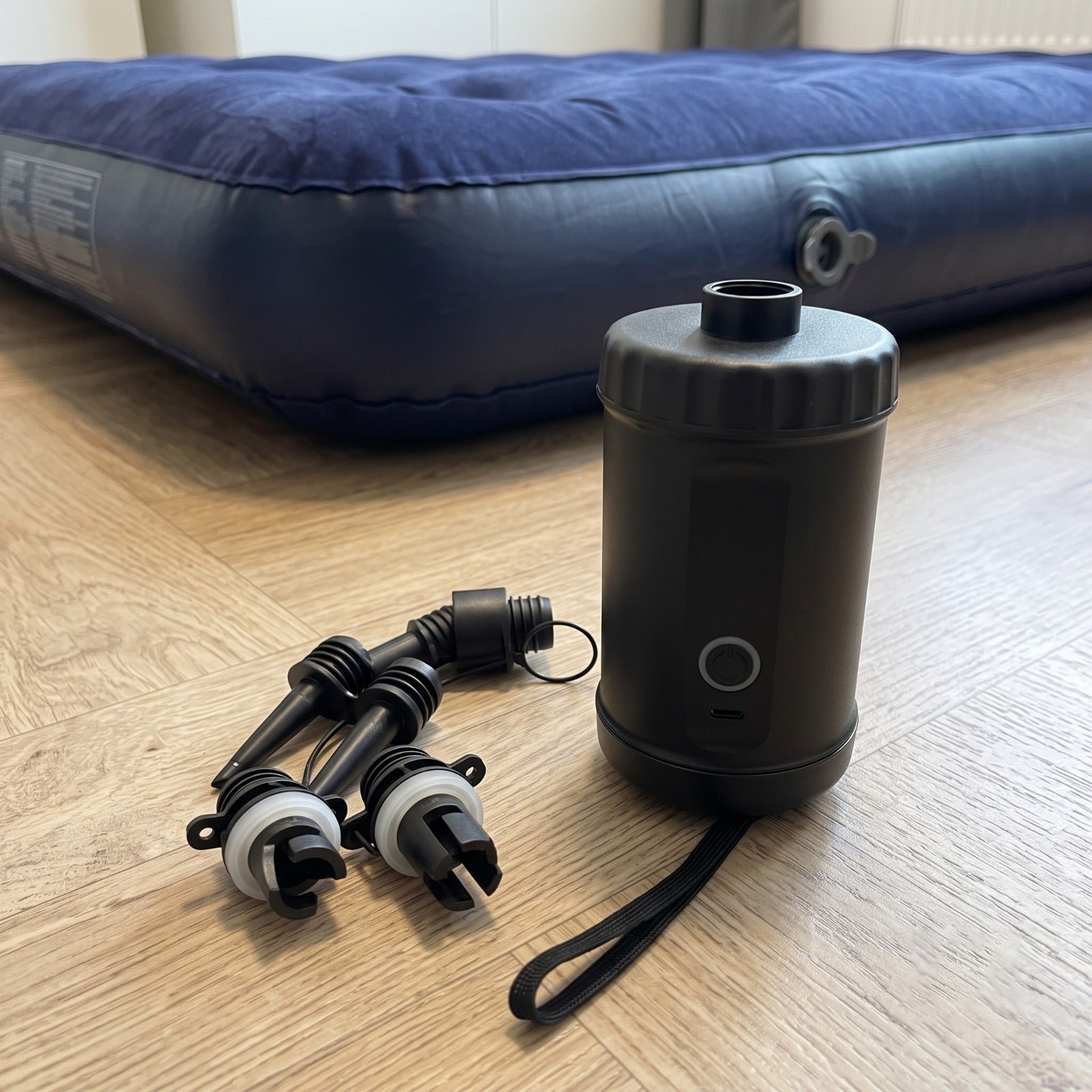 An Automatic Inflatable Air Pump and two air valves placed on a light wood floor in front of an inflated blue mattress.