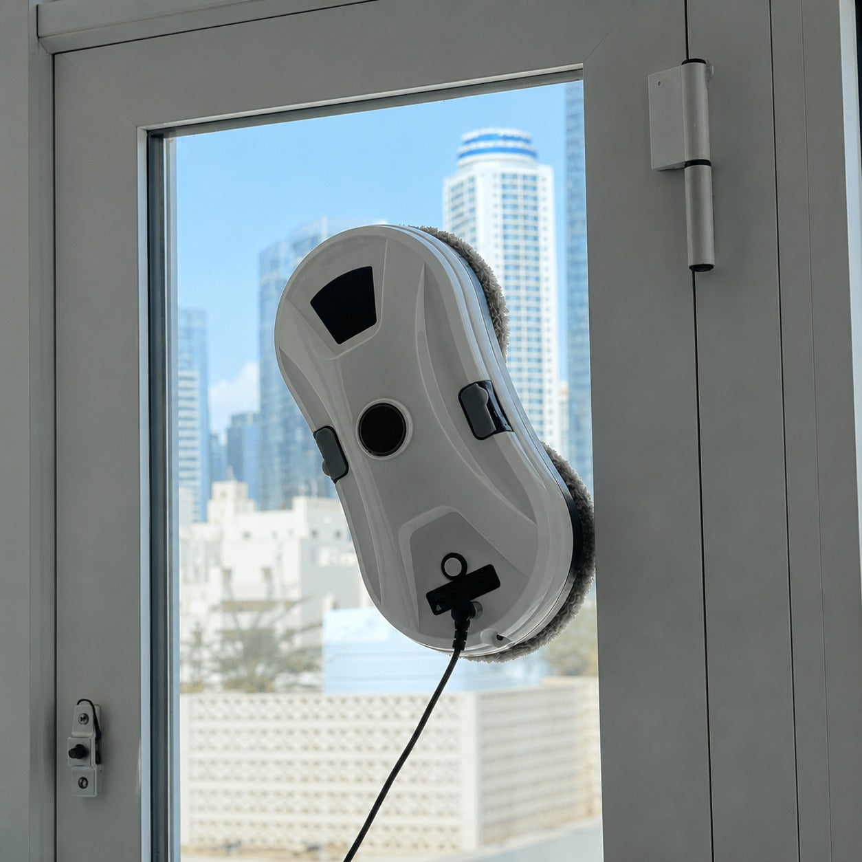 A white and gray electric window cleaning robot with a black cord is attached to a glass window of a gray door, with a city skyline visible in the background.