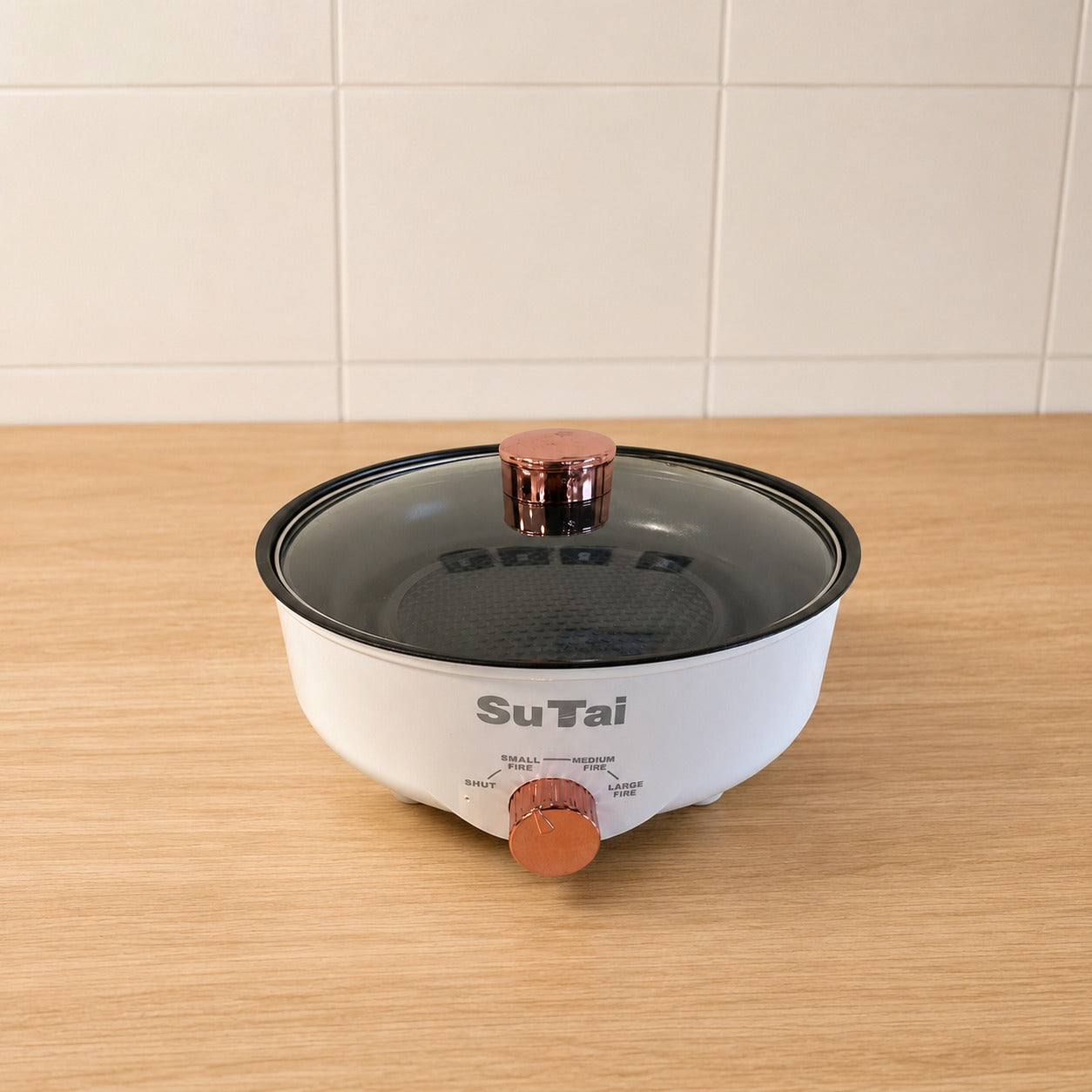 A Electric Frying Pan with a clear glass lid and copper-colored knobs, sitting on a light wood countertop against a white tiled wall.