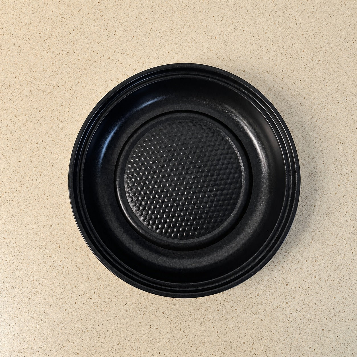 A black round Electric Frying Pan with a textured non-stick surface and raised rim, photographed from directly above on a light beige speckled countertop.