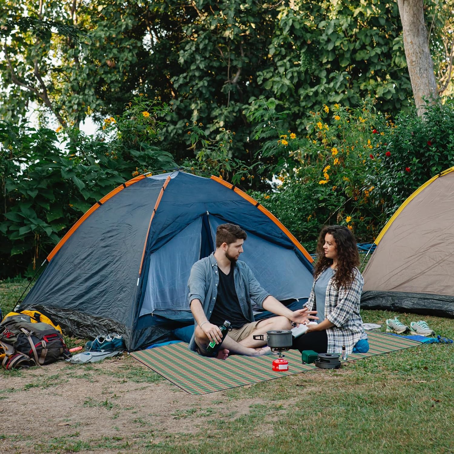 Two people sitting on a Foldable Waterproof Picnic Mat in front of a tent in a forested area.