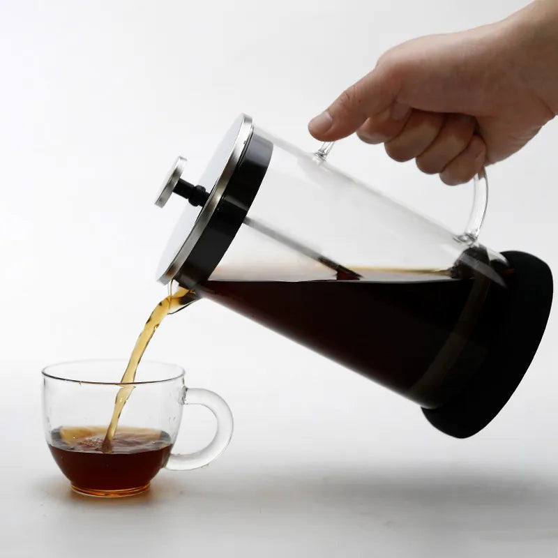 A hand pouring dark coffee from a clear glass French Press with a black base into a glass mug on a white surface.