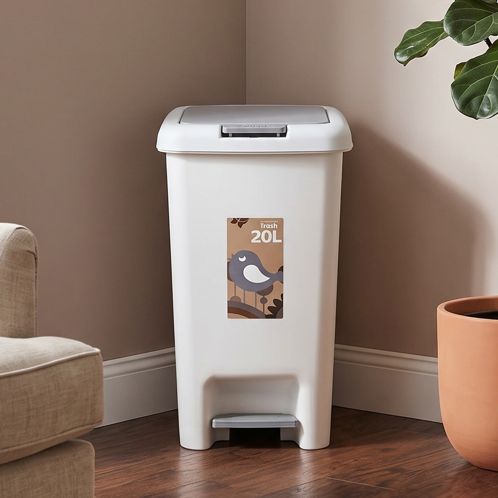 A 20L Pedal Trash Bin with a foot pedal, shown in a corner of a room with beige walls, a wooden floor, a partial beige armchair on the left, and a potted plant on the right.