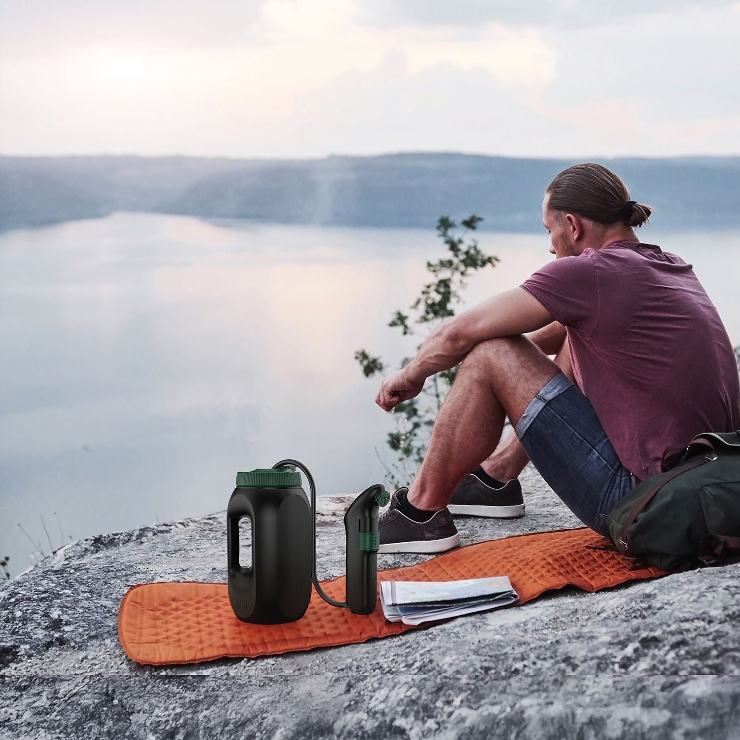 Man sitting outdoors on a cliffside during a camping trip with a Green Lion Bidet Pro portable hygiene device and large water tank beside him.