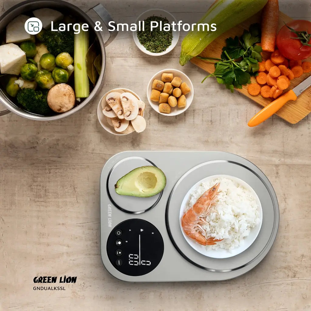 Green Lion Dual Kitchen Scale measuring food portions with large and small platforms, digital display, surrounded by fresh vegetables and cooked ingredients on a kitchen countertop.