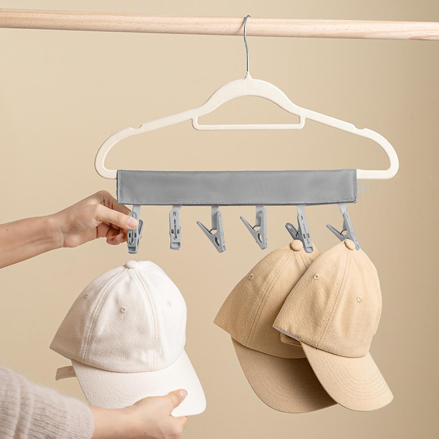 A person using a multi-clip hanger to hang baseball caps. The hanger is covered with a grey fabric sleeve and features several clips for organizing or drying hats efficiently.