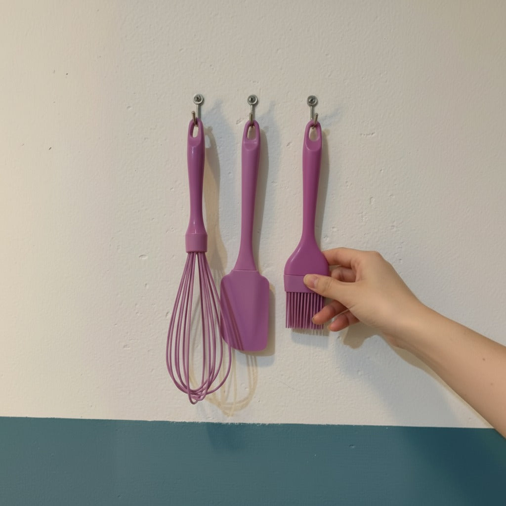 A Person is reaching for a brush, hanging on the wall beside a whisk and a spatula.