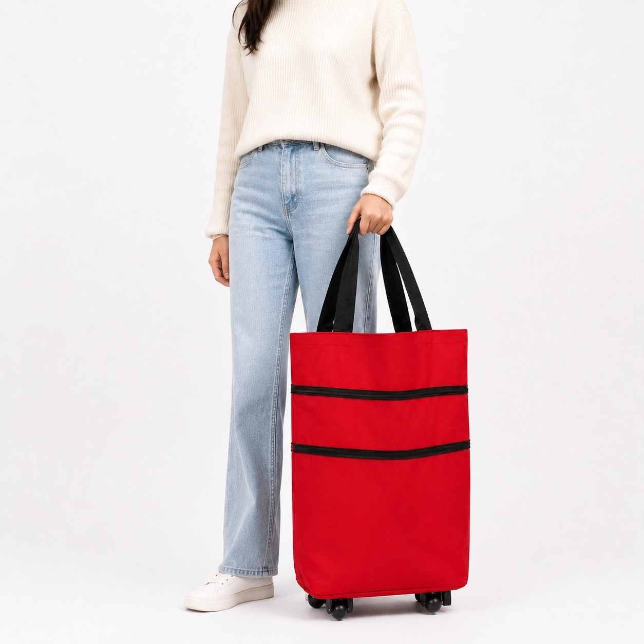 A person wearing a cream sweater, light blue jeans, and white sneakers holds a bright red rolling Shopping Trolley Bag gwith black handles and black horizontal trim against a clean white studio background.