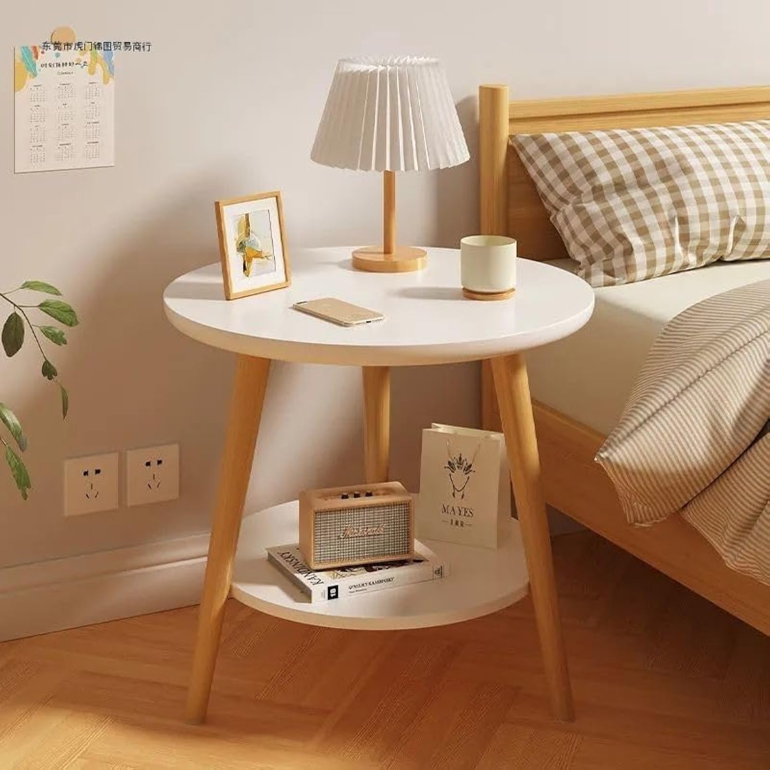 2-tier side table in a bedroom setting, with a cozy, minimal vibe. The top tier holds a frame, a table lamp , a cup, and a smartphone lying flat. On the bottom tier, there’s a vintage wooden radio and some books.