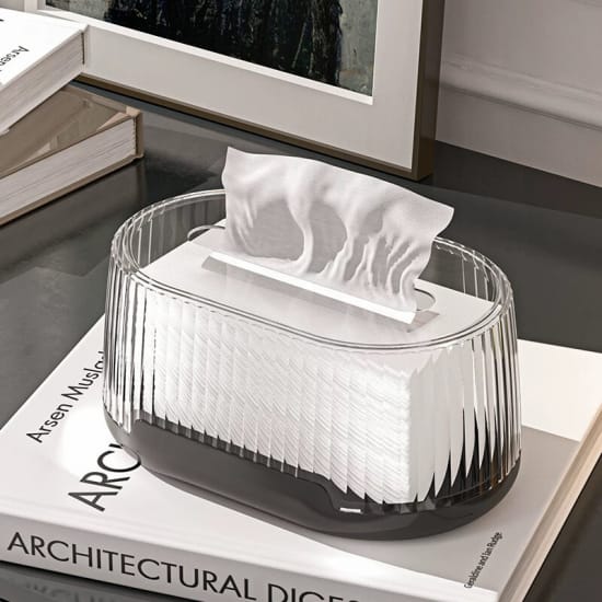 Oval shaped transparent tissue box is placed on top of books on a table, with one tissue pulled up and ready for use.