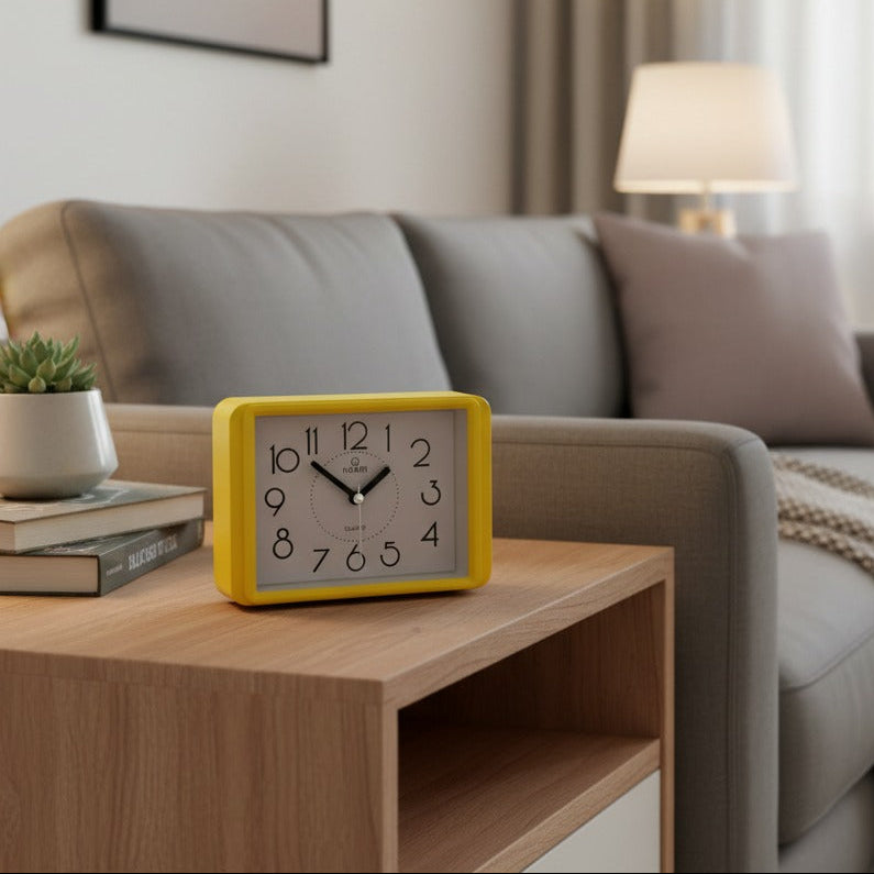 Rectangular alarm clock placed on a wooden coffee table in a modern living room with sofa and plants in the background.