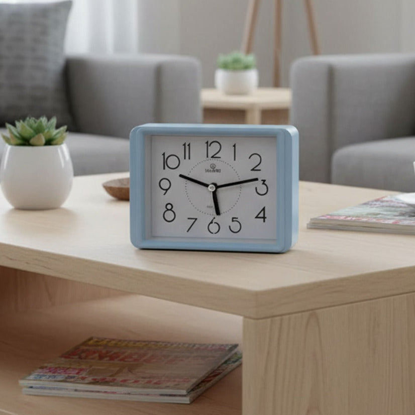 Rectangular blue alarm clock placed on a wooden coffee table in a modern living room with sofa and plants in the background.