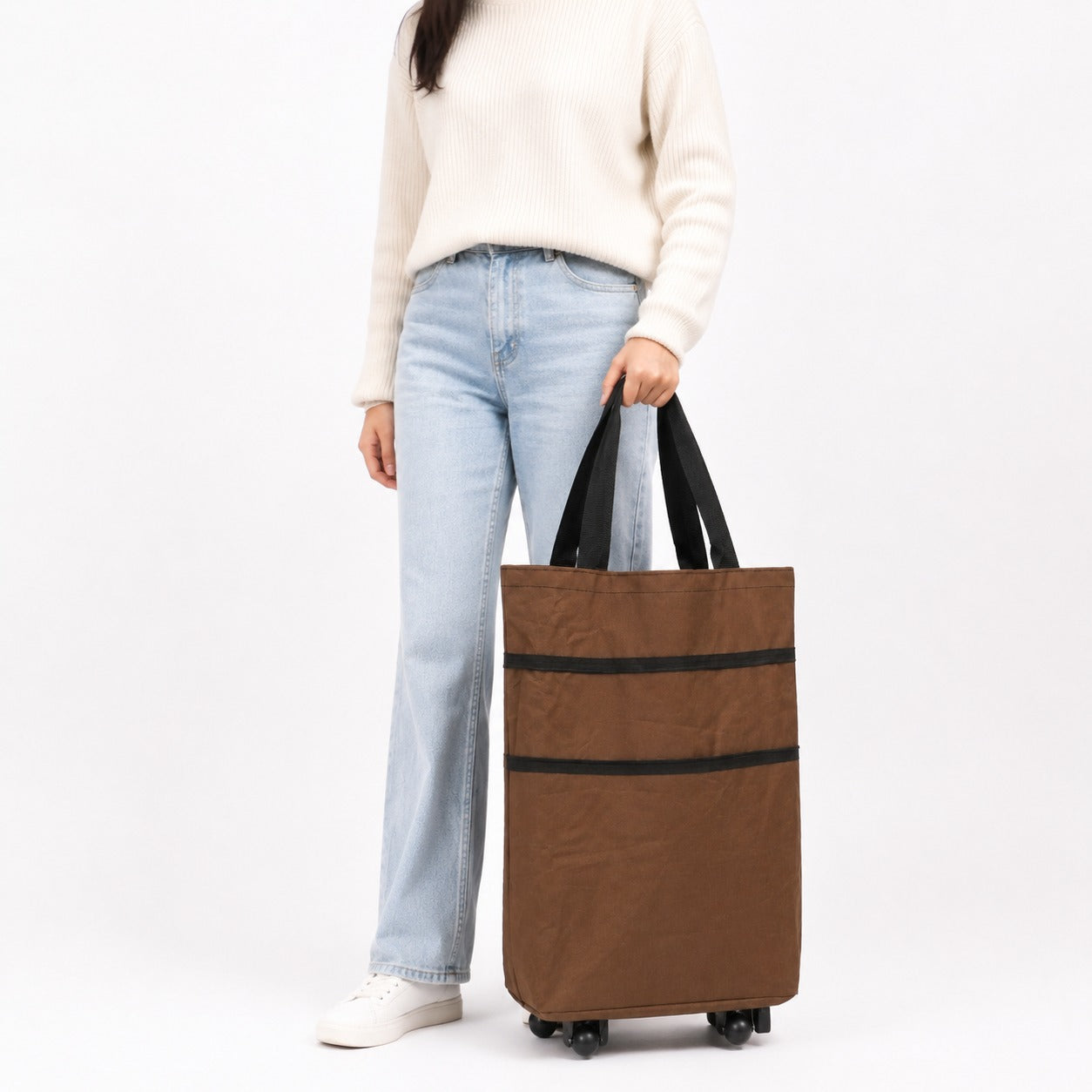 A person wearing a cream sweater, light blue jeans, and white sneakers holds a brown Shopping Trolley Bag with black handles and black horizontal stripes against a plain white studio background.