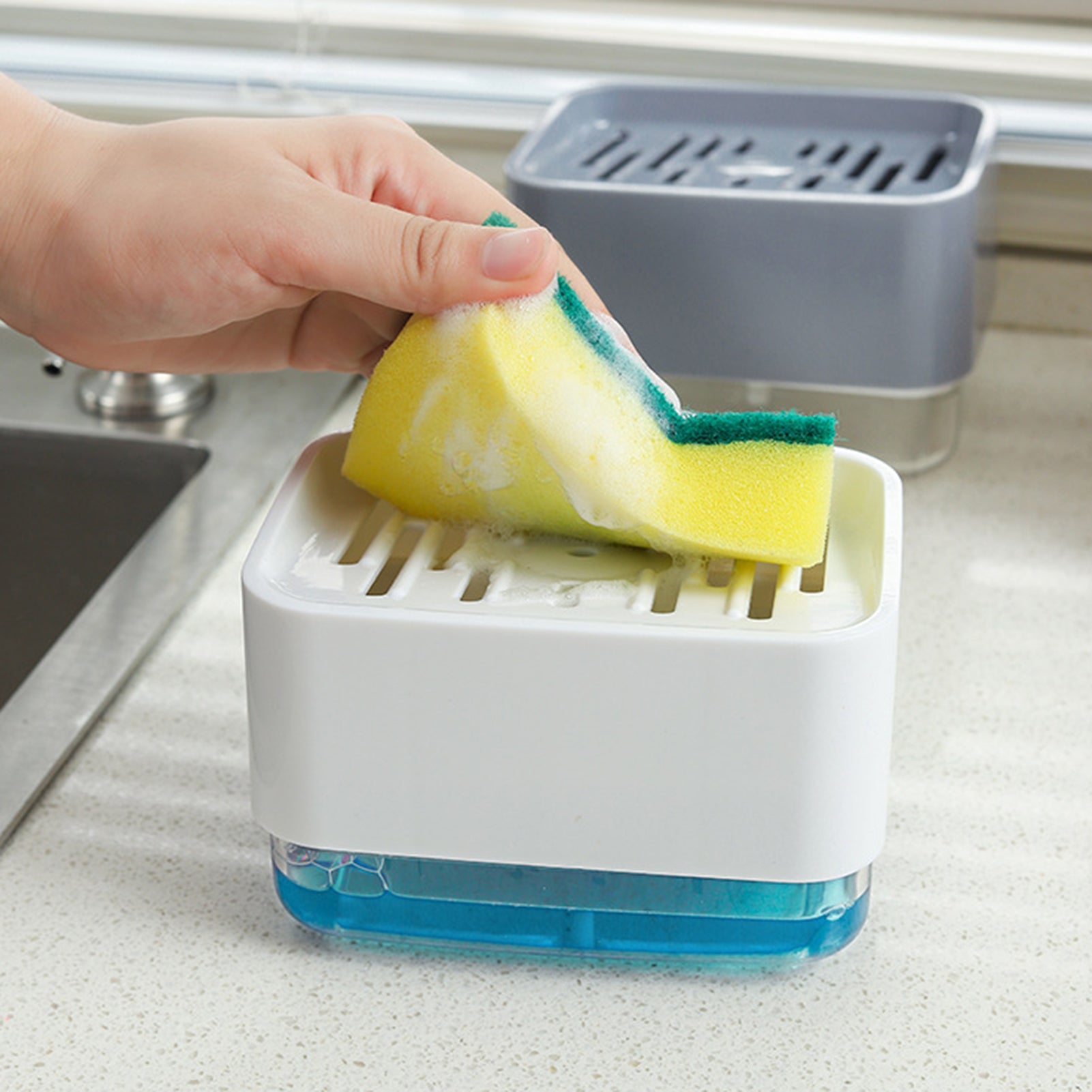A compact sponge holder with a built-in soap dispenser, shown being pressed with a sponge to release liquid soap, placed beside a kitchen sink for easy dishwashing.