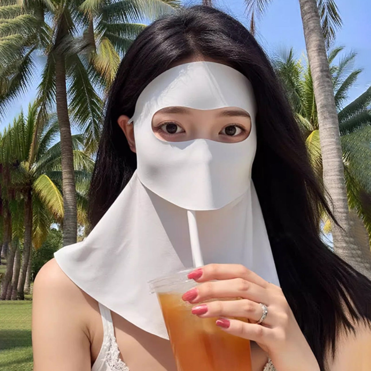 Woman Wearing sun mask, sipping iced tea without removing the mask . Palm trees behind.