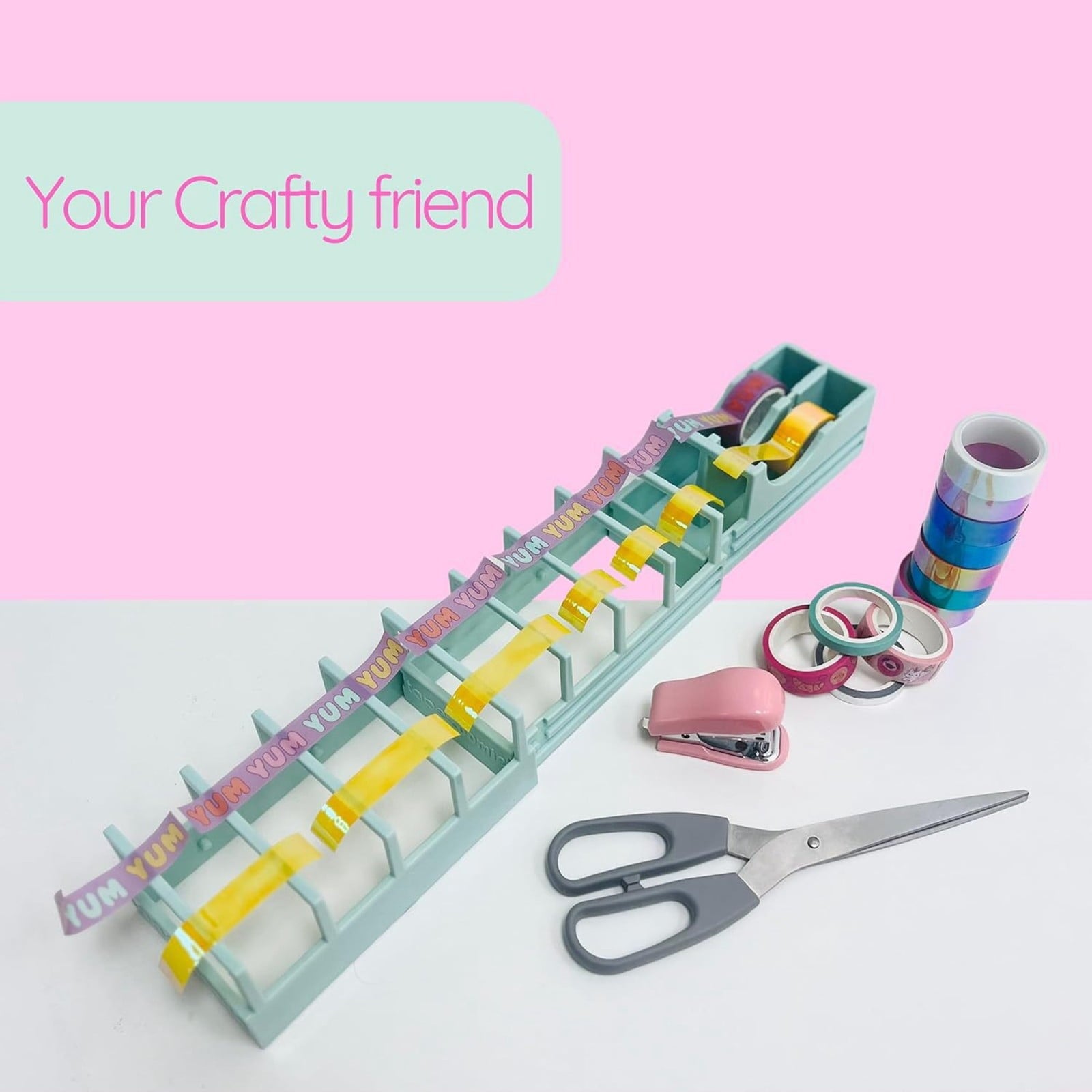 A colorful crafting setup with decorative tapes organized in a dispenser, scissors, a pink stapler, and stacked washi tape rolls