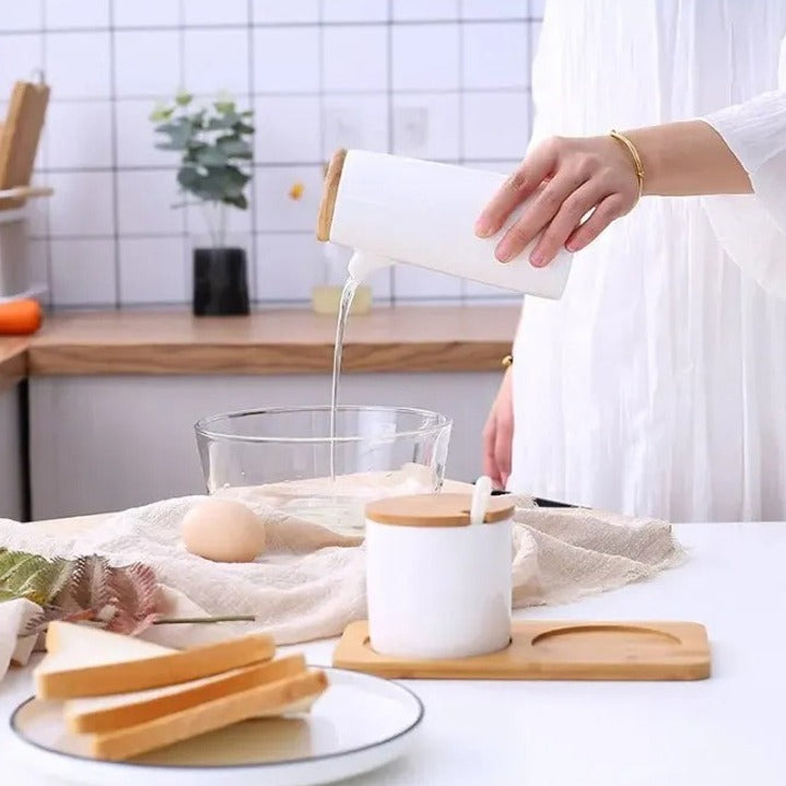 A person pouring liquid from a ceramic bottle into a bowl in a kitchen, with a matching seasoning container set placed on a wooden stand nearby.