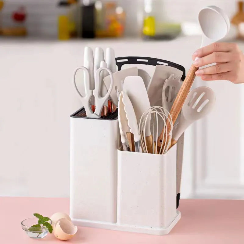 A person is taking from 18-piece silicone kitchenware set in a  holder including spatulas, ladle, tongs, whisk, pasta server, brushes, and scissors, neatly organized on a kitchen countertop.