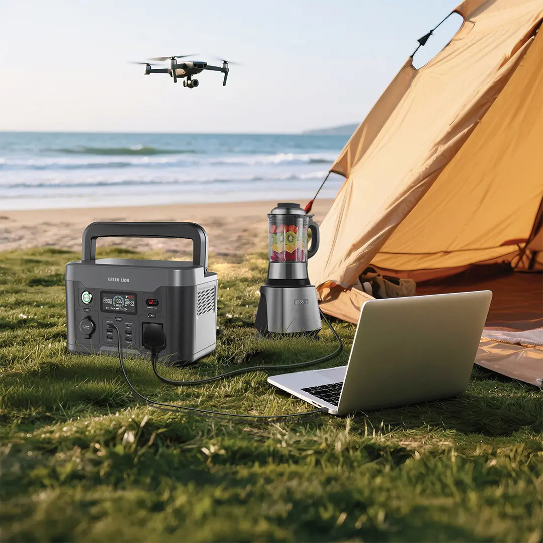 A Green Lion power station is placed on the grass beside a beachside camping setup. It is connected to a laptop and a blender, both powered simultaneously, showing its multi-device capability.
