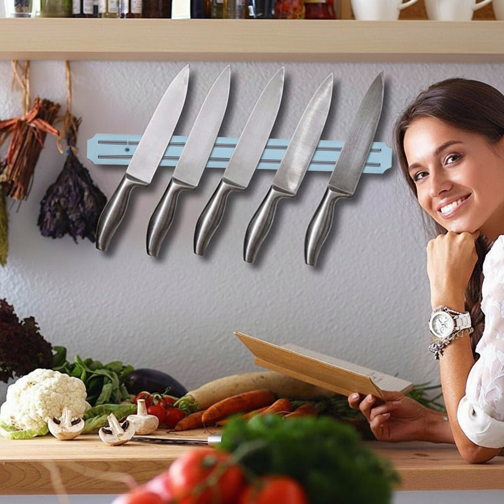 A wall-mounted magnetic strip holding multiple kitchen knives neatly in a row, placed above a kitchen counter where vegetables are prepared, with a person sitting nearby.