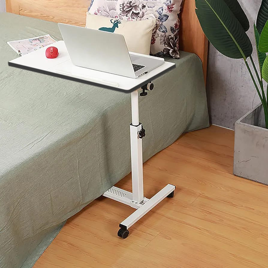 White Adjustable Overbed Laptop Stand Table with a laptop on top, positioned on the floor beside a Bed.