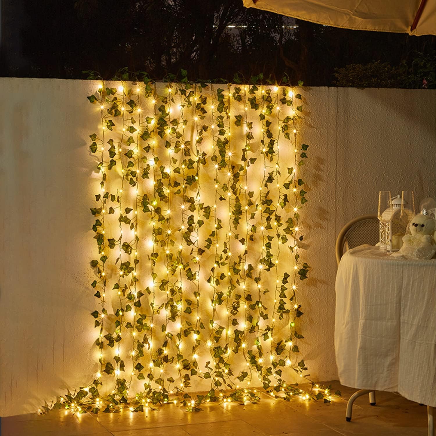 Decorative wall with green foliage and string lights, next to a table with a white tablecloth and decorative items.