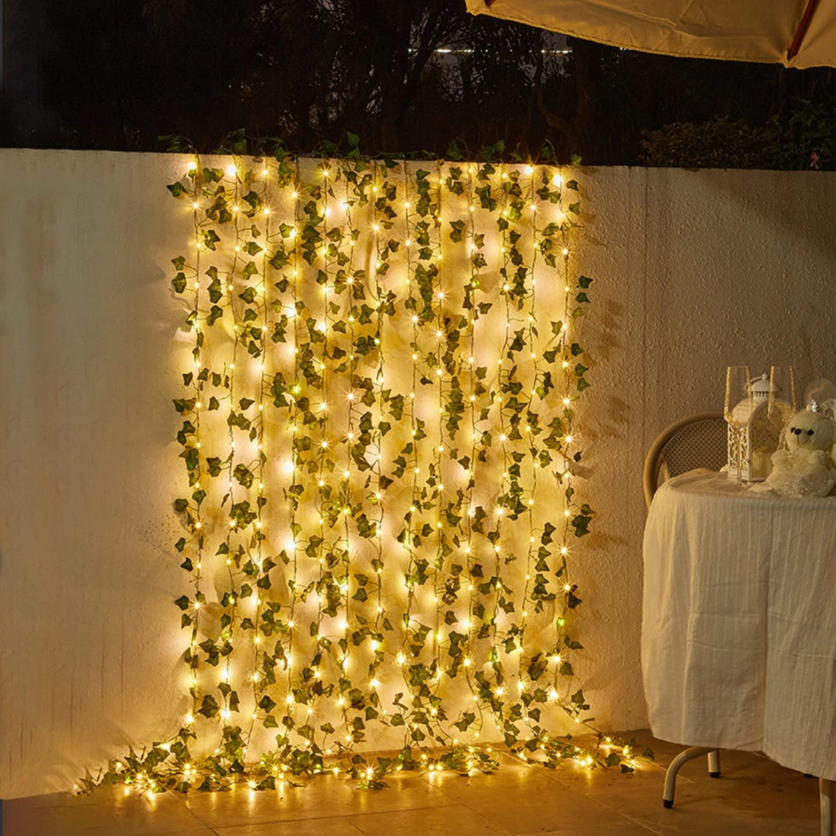 Decorative wall with green foliage and string lights, next to a table with a white tablecloth and decorative items.