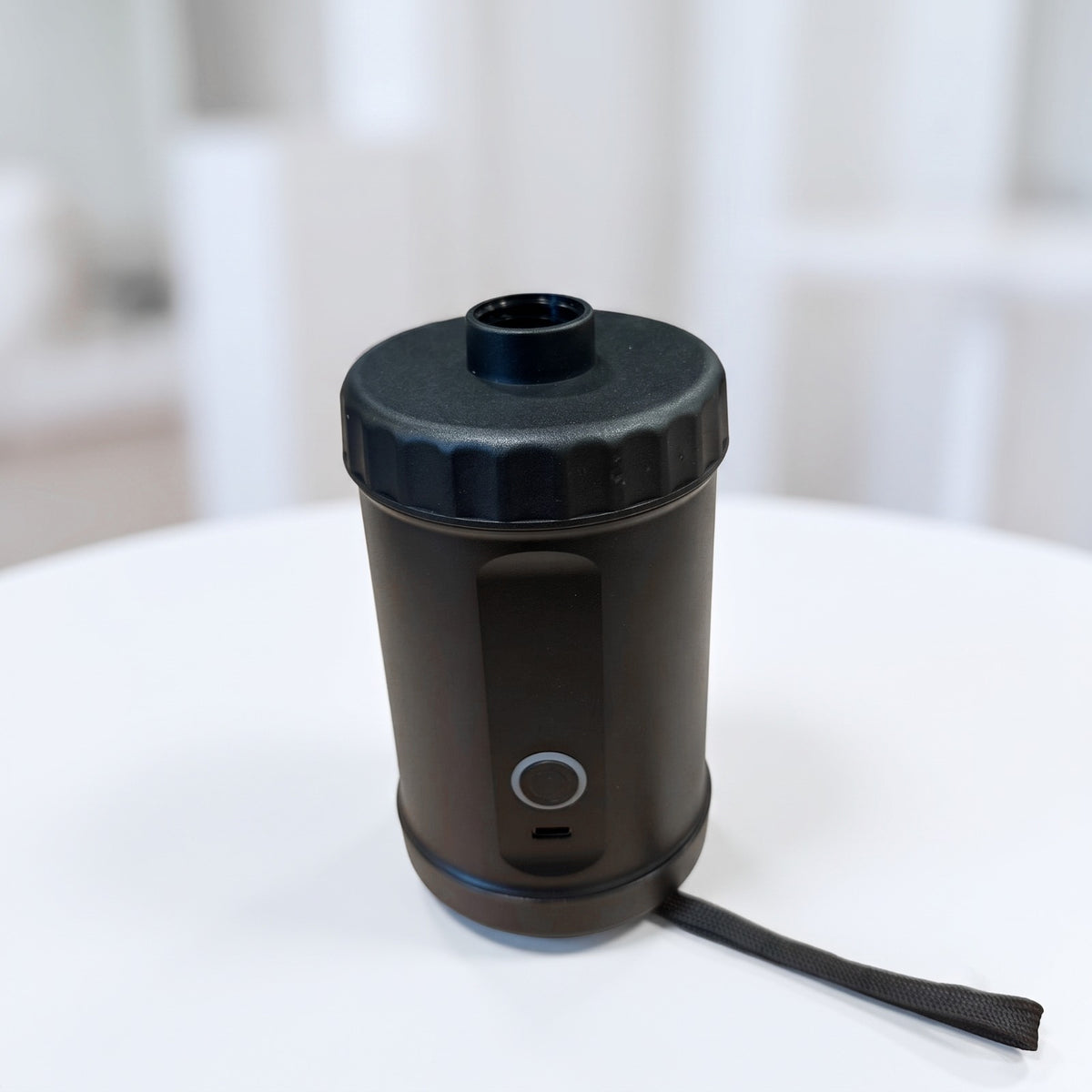 A black Automatic Inflatable Air Pump with a ribbed screw-on lid and a small carrying strap, photographed on a white tabletop with a softly blurred indoor background.
