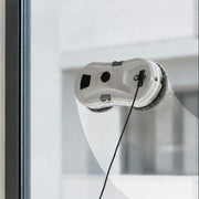 A white and gray window cleaning robot with a black power cord, shown attached to a glass window in a modern interior setting.