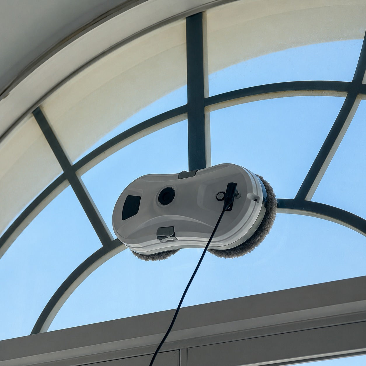 A window cleaning robot with circular brushes and a black power cord, shown attached to a glass window under a large arched roof structure.