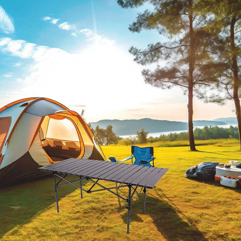 Outdoor camping scene with a dome tent pitched on grass beside a folding camp table, blue folding chair, and a small cooler, with trees and a lake in the background under a bright sky.