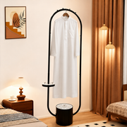 A Cloth Incense Burner Stand with an arched top stands in a bedroom setting, holding a white long-sleeve tunic. The room includes warm brown curtains, hanging pendant lights, a bedside table with a lamp, and a framed wall art piece.