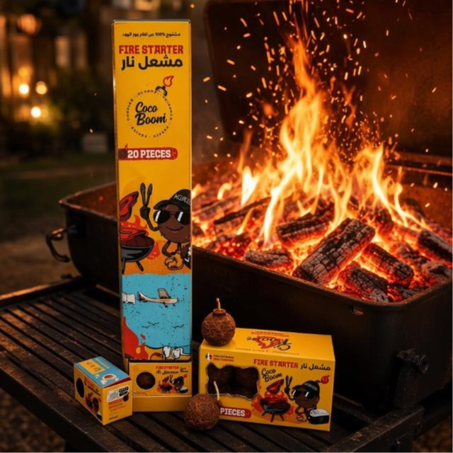 Coco Boom Coconut Shell Fire Starter and their packaging displayed on a grill table beside an active outdoor fire. The yellow boxes show cartoon graphics and the text 'FIRE STARTER' and '20 PIECES', with several brown fire starter balls placed in front.