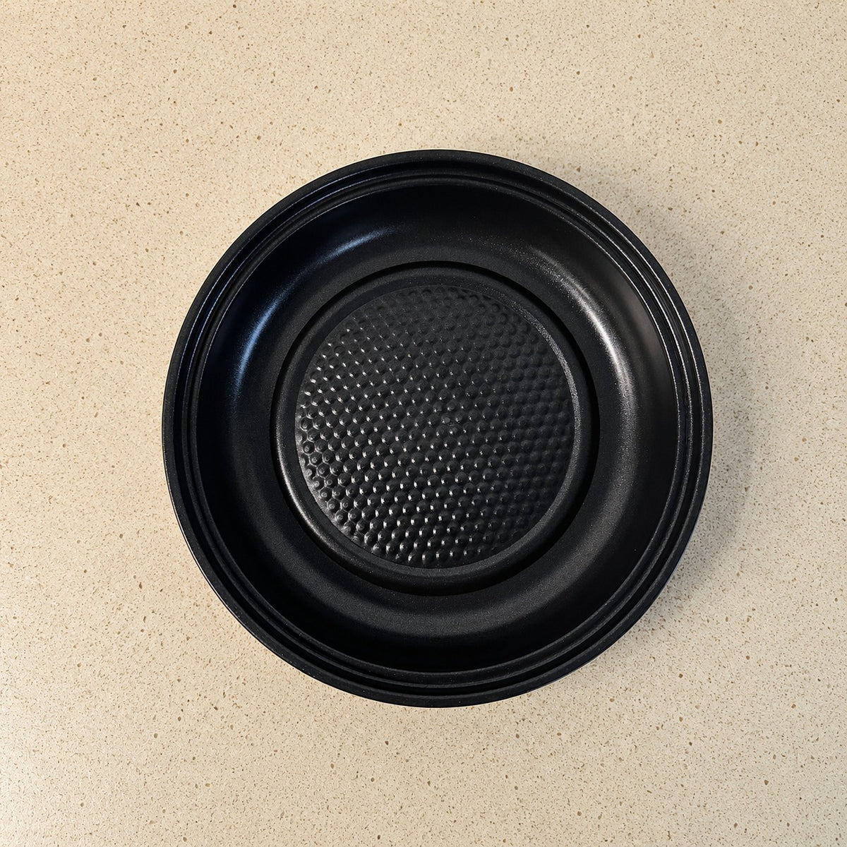 A black round Electric Frying Pan with a textured non-stick surface and raised rim, photographed from directly above on a light beige speckled countertop.