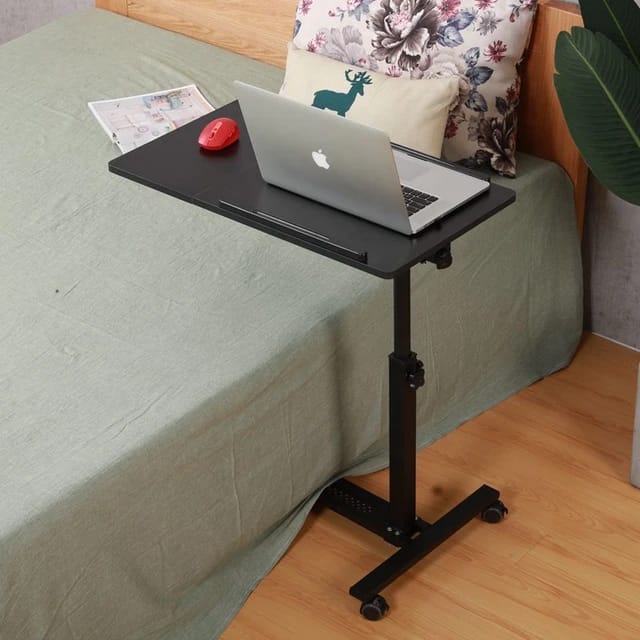 Black Adjustable Overbed Laptop Stand Table with a laptop on top, positioned on the floor next to a bed