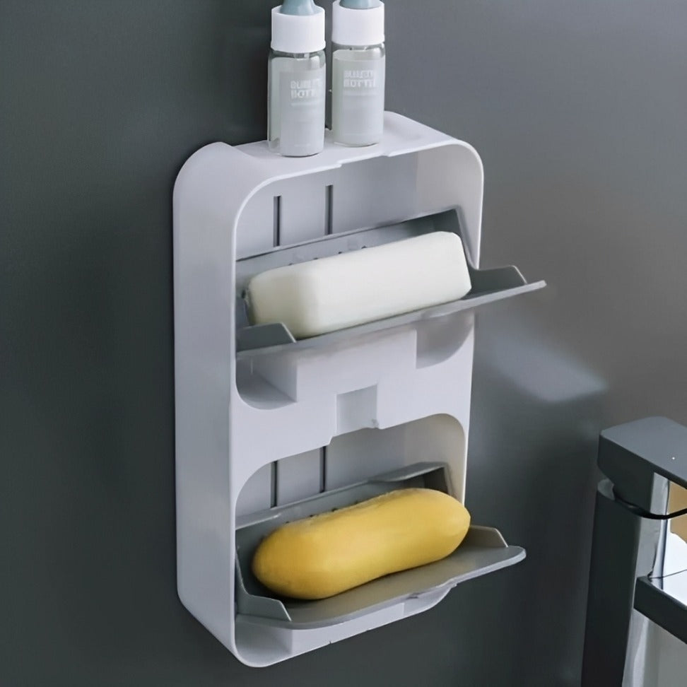 A wall-mounted soap holder with two slanted trays. A white soap bar is placed on the upper tray, and a yellow soap bar on the lower tray. On top of the holder, there are two small bottles