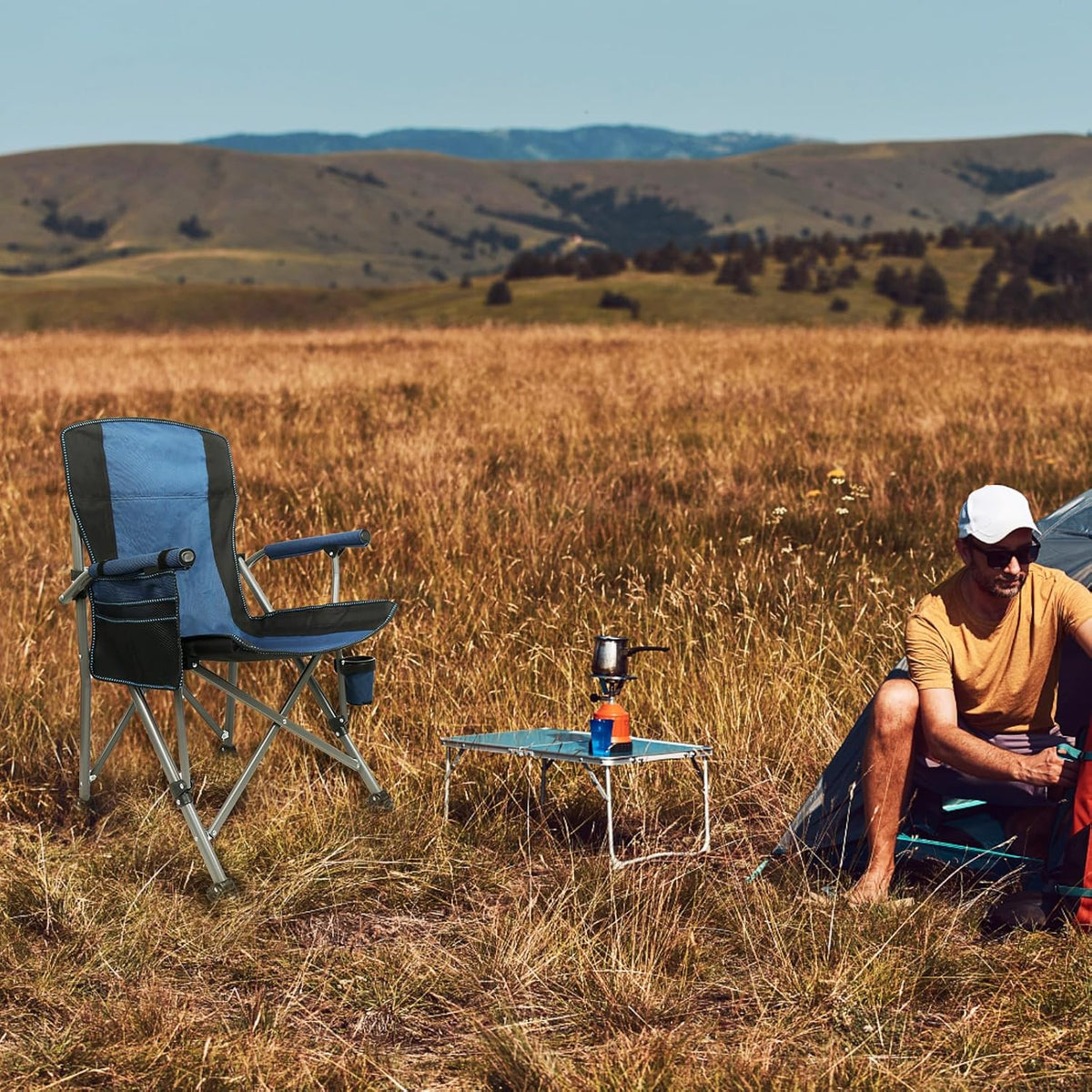 Person sitting in a field with camping chair and table, surrounded by nature.