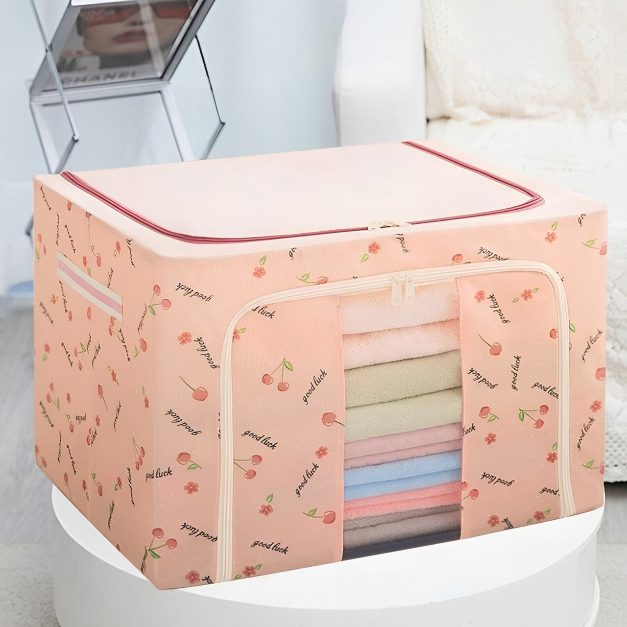 A pink Foldable Cloth Storage Organizer Box with a cherry print pattern, shown on a white pedestal in a bright indoor setting. The box has a zippered top and a front window displaying folded towels or blankets in pastel colors.