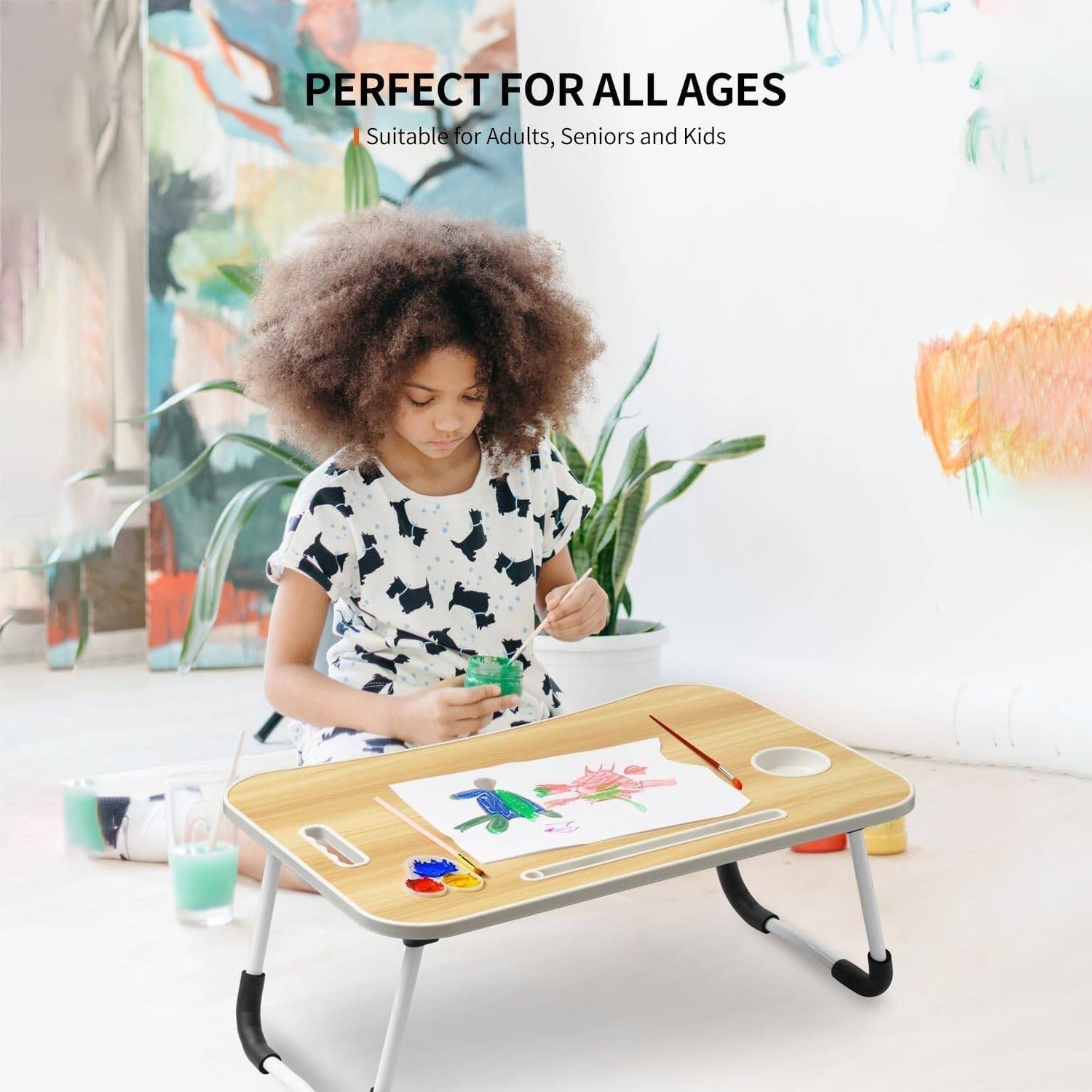 A young girl with an afro sits at a small folding Foldable Laptop Table with a tray table, drawing on a white sheet of paper. The scene is set in a bright, modern room with a plant and colorful abstract art in the background.