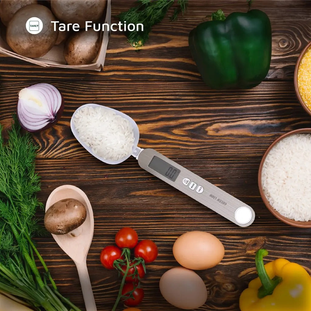 A digital measuring spoon scale with a Tare function, filled with rice, is placed on a wooden kitchen table surrounded by fresh vegetables and ingredients like eggs, tomato, mushrooms, and bell pepper.