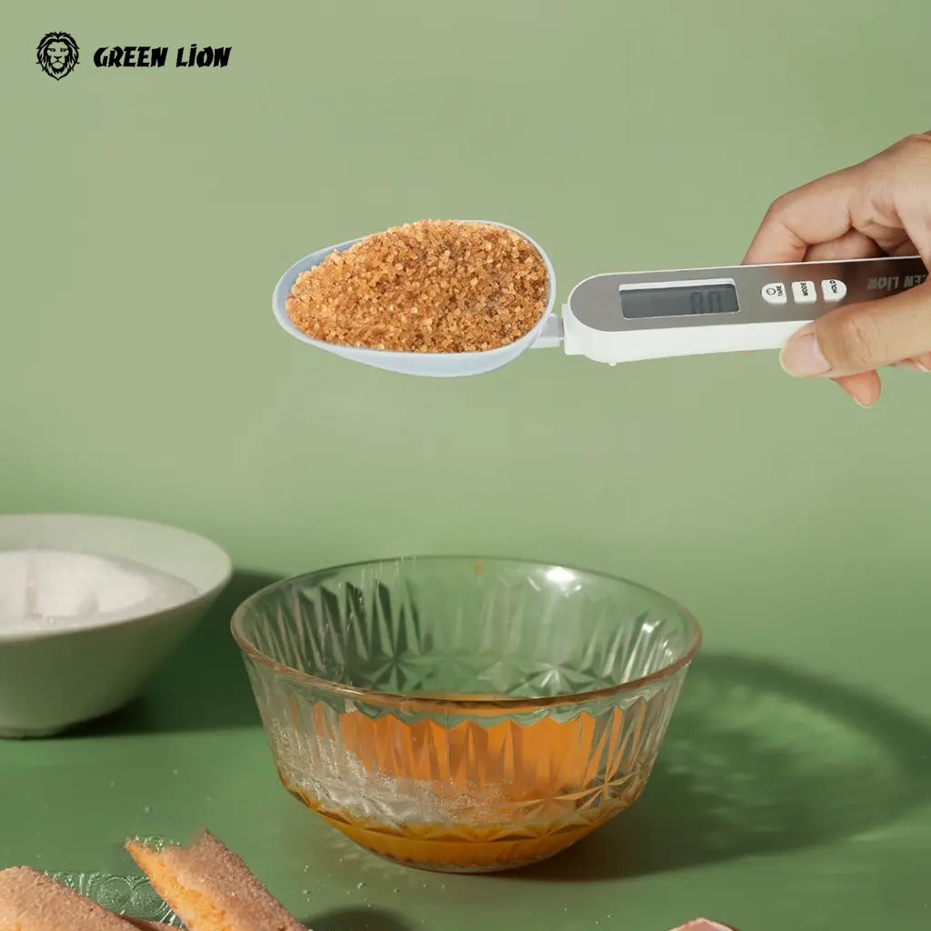 A digital measuring spoon scale branded Green Lion, held by a hand, measuring brown sugar above a glass bowl on a green background.