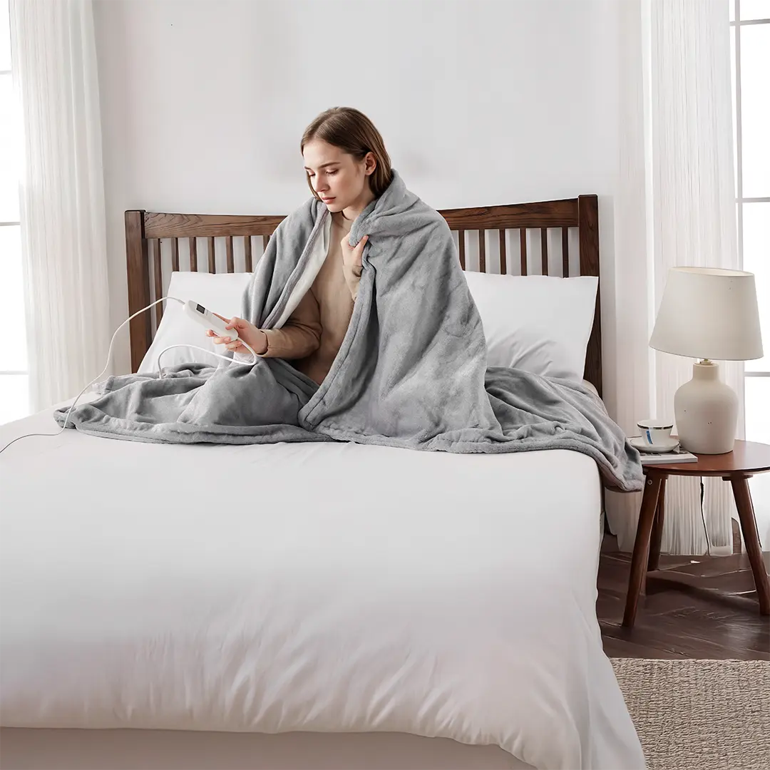 A woman sitting on a bed wrapped in a gray Green Lion Electric Blanket with Heating Function, using a Handheld Remote Controller in a bright bedroom.