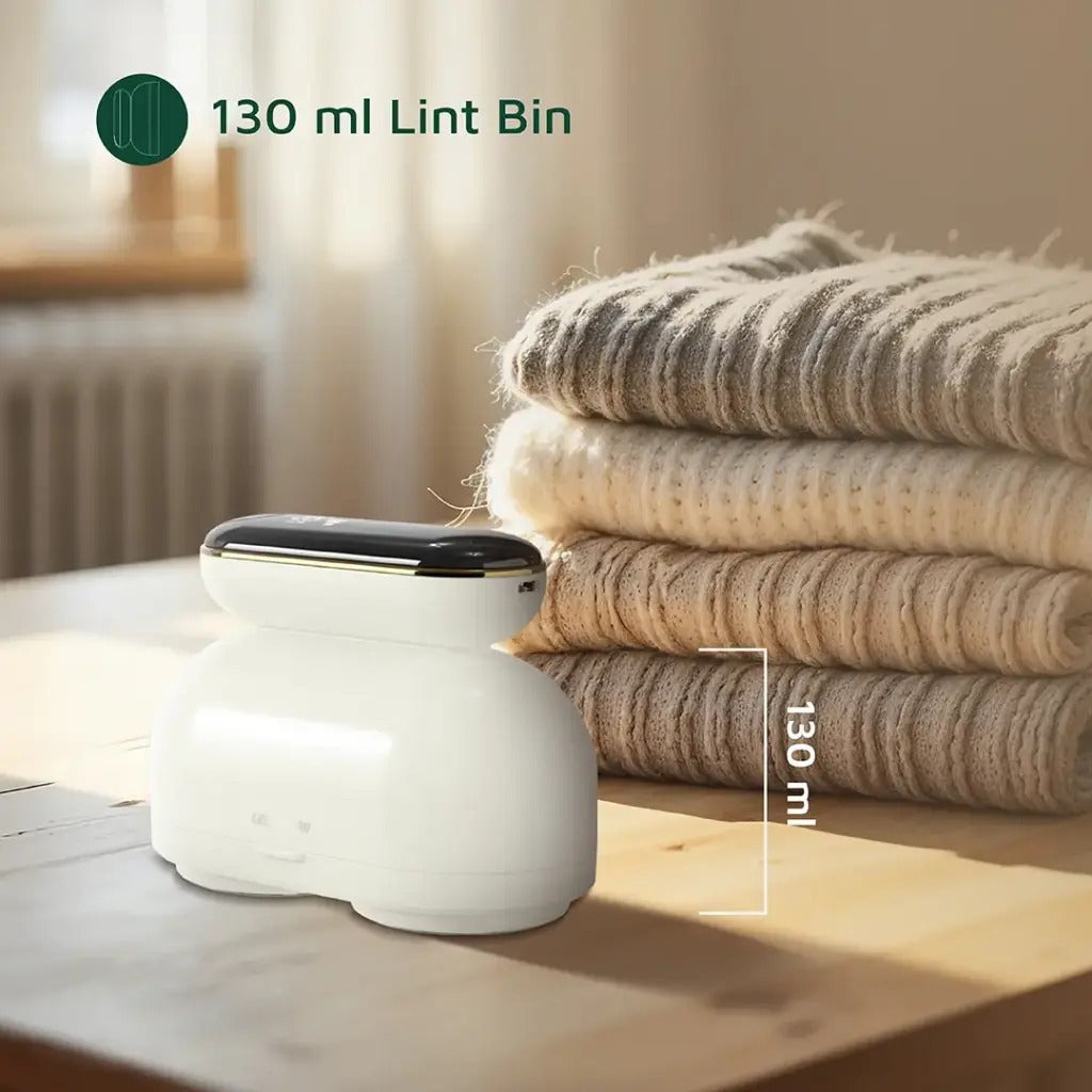 Green Lion DualPro Digital Fabric Remover placed on a wooden surface beside a neatly folded stack of sweaters. The image highlights its 130 ml lint bin capacity, emphasizing efficiency and storage convenience. Warm lighting adds a cozy, home-like atmosphere.