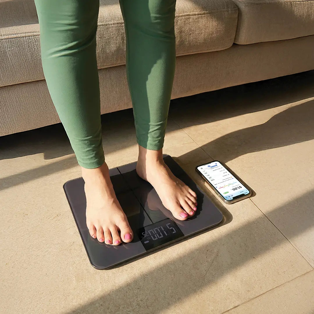 Person standing on a Green Lion  Smart Body Scale with a smartphone displaying app on a beige floor.