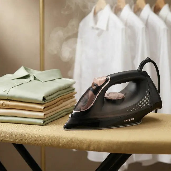 Black Green Lion Steam Iron on an ironing board with folded shirts and hanging clothes in the background