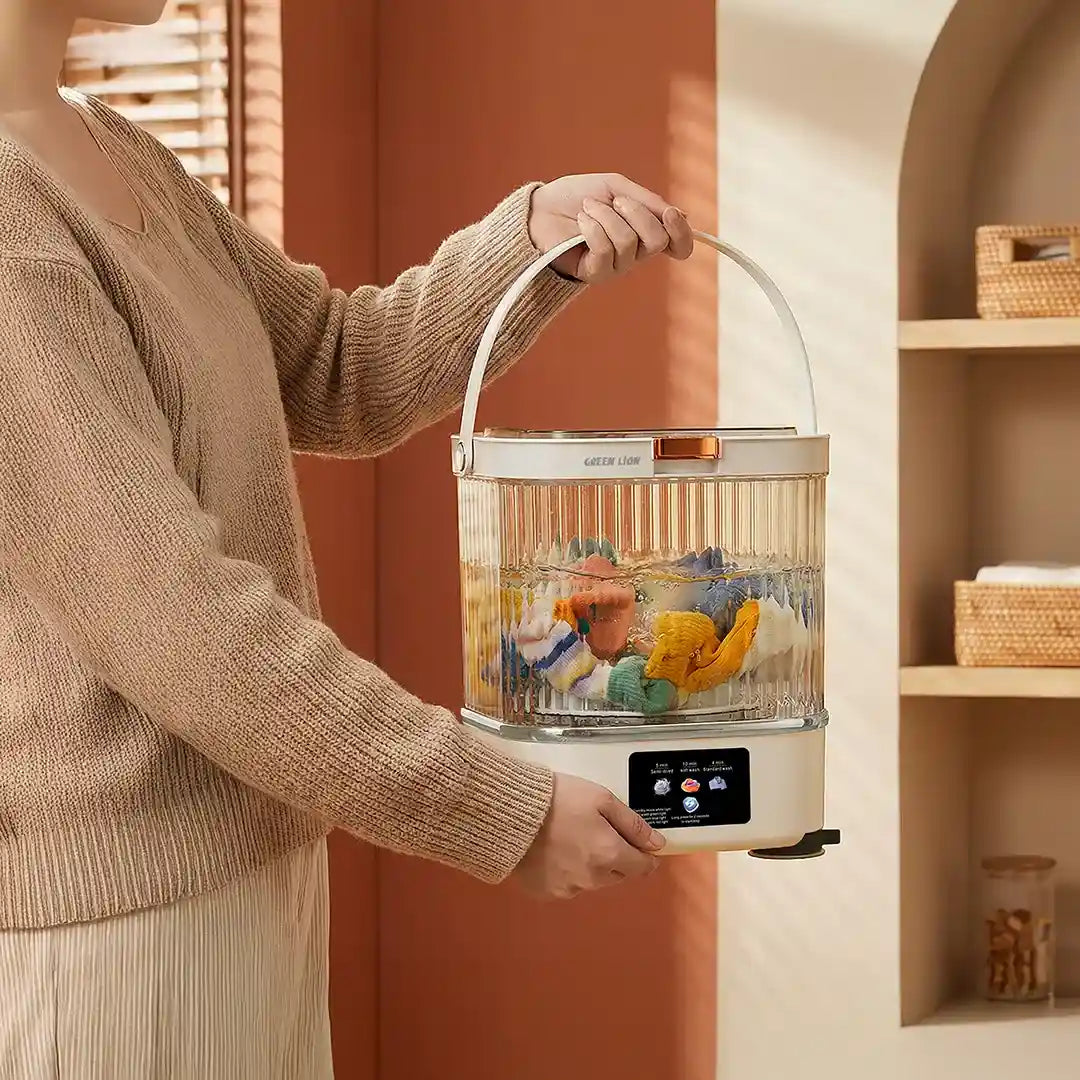 Person holding a Green Lion 12L Portable Mini Washing Machine with clothes inside against a warm-toned wall.