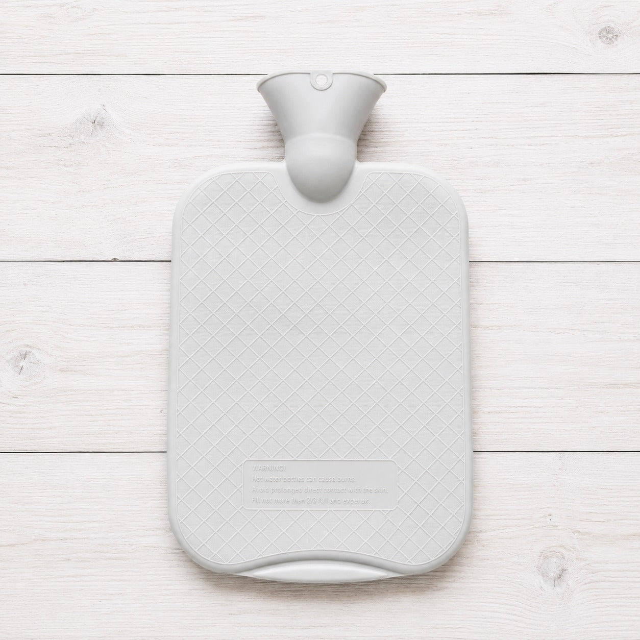 A light gray hot water bottle with a textured diamond-patterned body and a matching gray neck, shown from a top-down view on a white wooden plank background.