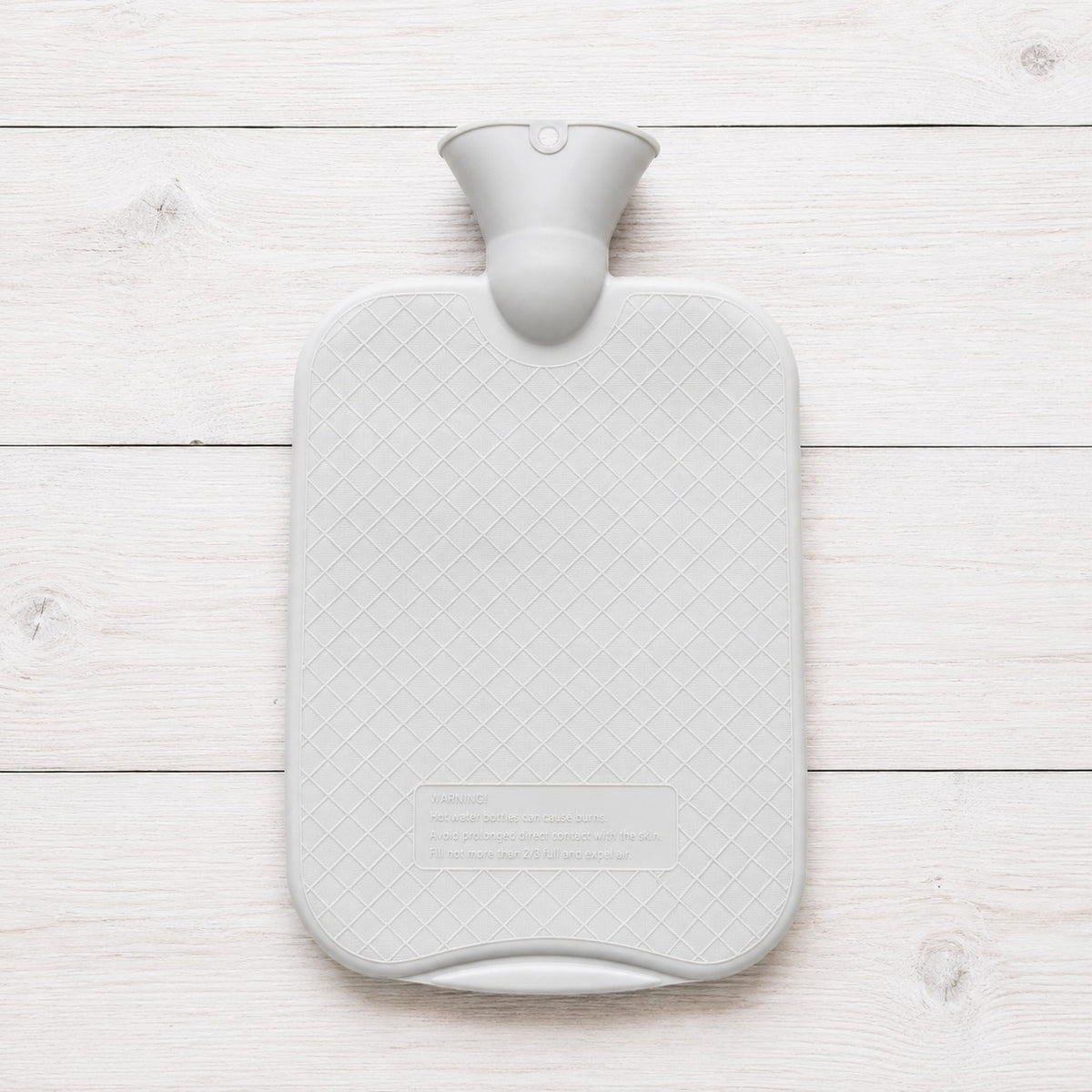 A light gray hot water bottle with a textured diamond-patterned body and a matching gray neck, shown from a top-down view on a white wooden plank background.