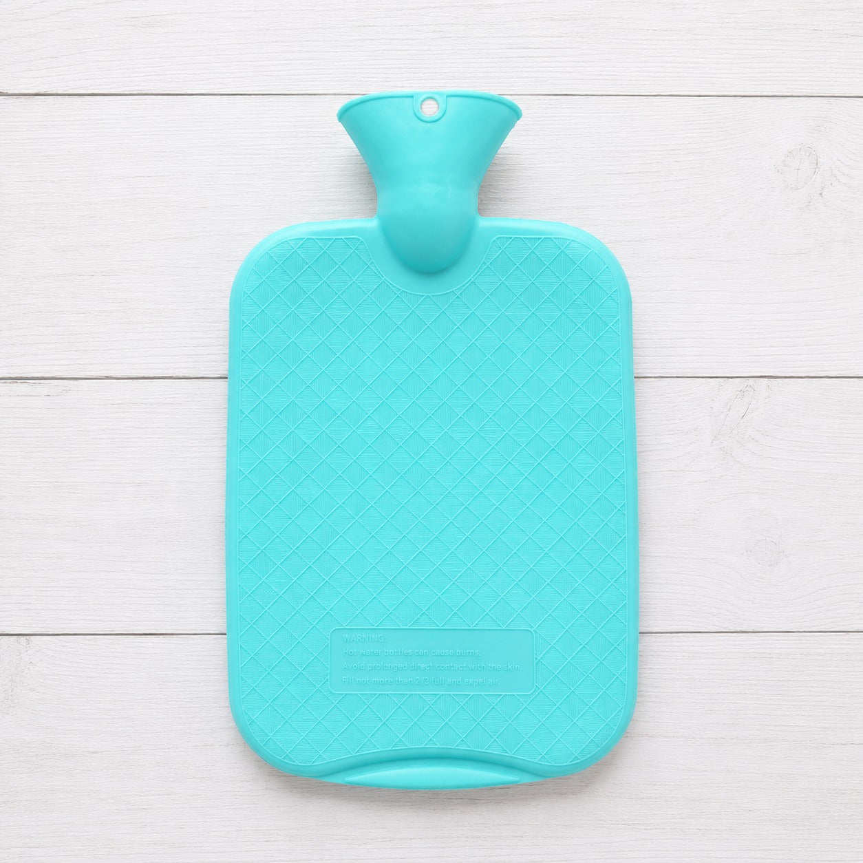 A hot water bottle with a diamond-patterned textured surface, shown from a top-down view on a white wooden plank background.