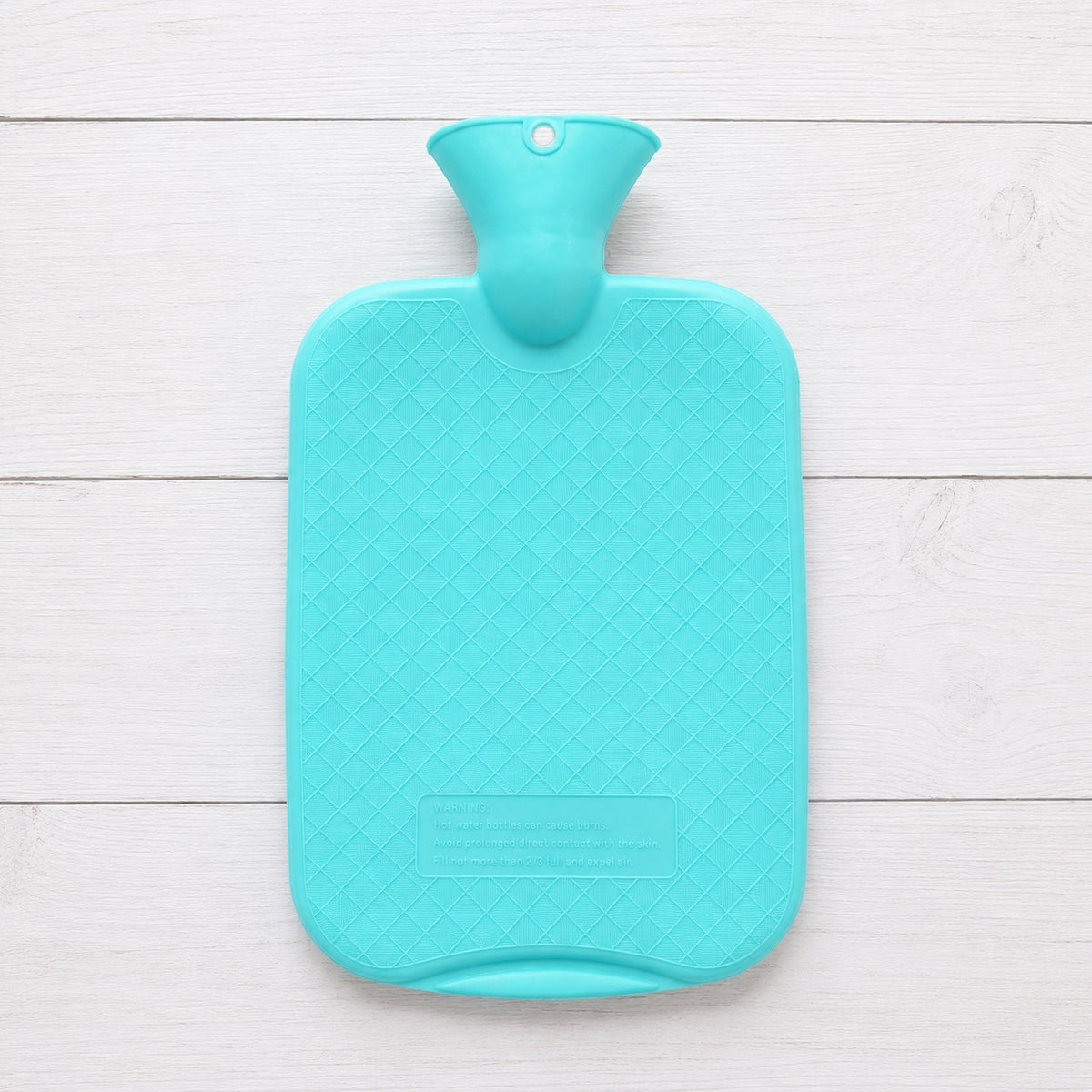 A hot water bottle with a diamond-patterned textured surface, shown from a top-down view on a white wooden plank background.